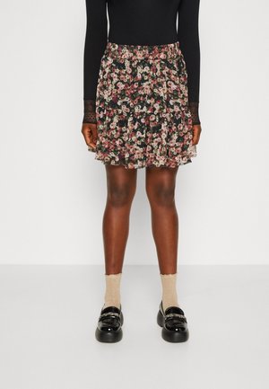 Femme portant un haut noir à manches longues, une jupe courte à motif floral, des chaussettes beige et des mocassins noirs brillants, debout devant un fond blanc.