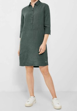 Femme portant une robe en lin verte foncée, longueur genou, boutonnée, à manches trois-quarts, avec des baskets blanches, debout devant un fond uni.