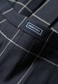 Tissu bleu marine avec un motif à carreaux, présentant des lignes blanches. L'étiquette indique "Superdry" sur un fond bleu foncé, située à la taille.