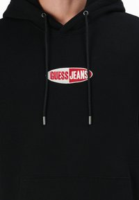 Juoda džemperis su kapuce, pagamintas iš medvilnės mišinio, su raudonu ir baltu siuvinėtu "GUESS JEANS" logotipu. Į komplektą įeina raišteliai ir priekis kišenė.