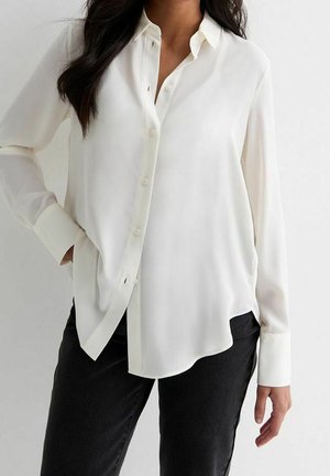 Kobieta ubrana w luźną białą koszulę na guziki z długimi rękawami i czarne spodnie, stojąca na tle jednolitego jasnego tła.