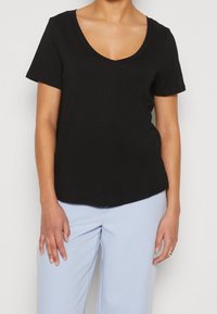 Kobieta w czarnej koszulce z krótkim rękawem i dekoltem w serek oraz jasnoniebieskich spodniach, stojąca z ramionami zrelaksowanymi wzdłuż ciała.