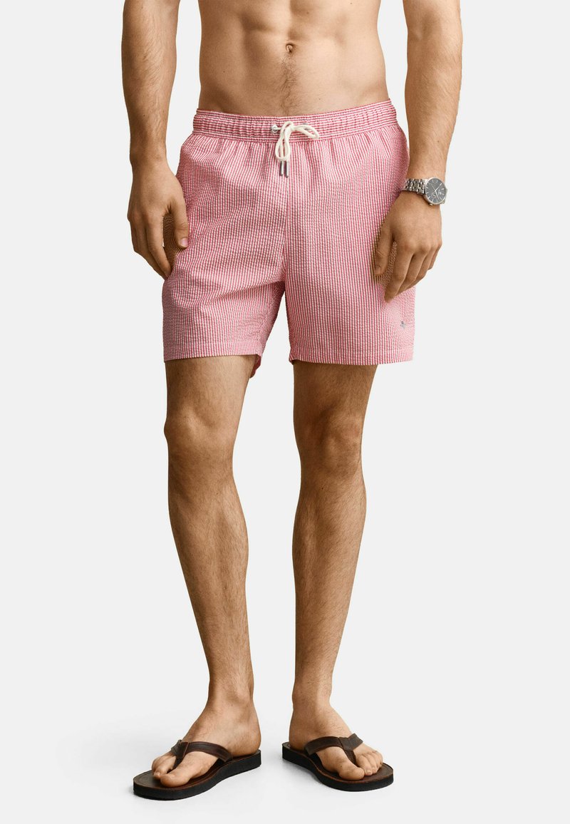 Homme portant un short de bain rayé rouge et blanc avec un cordon blanc, des tongs marron et une montre-bracelet argentée, debout sur un fond blanc.