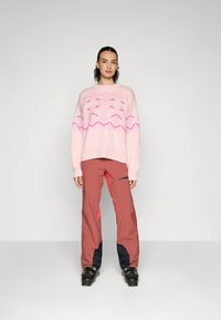 Jasnoróżowy dziergany sweter z geometrycznymi wzorami, zestawiony z czerwonymi i granatowymi spodniami narciarskimi. Model nosi czarne buty narciarskie.