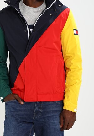 Mann trägt eine Color-Block-Jacke mit marineblauen, roten, gelben und grünen Abschnitten, kombiniert mit blauen Jeans und einem grauen T-Shirt darunter.