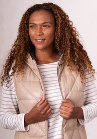 Woman with curly hair wearing beige quilted vest and striped long sleeve shirt, holding the vest collar and looking right.