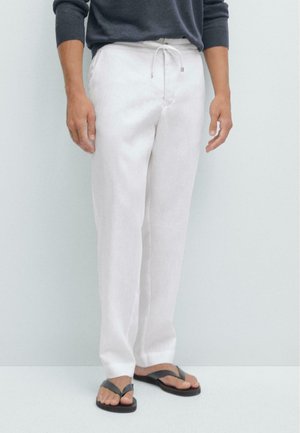 Homme portant un pantalon blanc à cordon et des tongs noires, debout devant un fond clair avec les mains détendues le long du corps.
