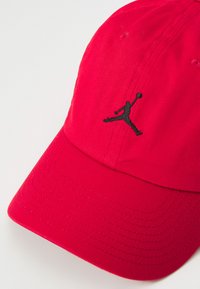 Sarkans cepure, izgatavota no teksturēta auduma, ar melnu izšuvumu priekšpusē, kurā redzams Jumpman logos. Ielocīta viziera ar izšūtām detaļām.