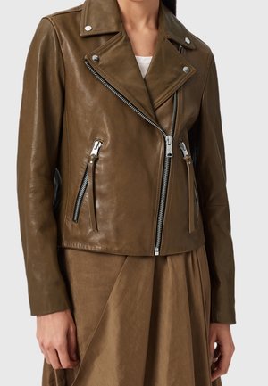 Femme portant une veste de biker en cuir vert olive avec des fermetures éclair argentées et des boutons-pression sur une jupe marron assortie.