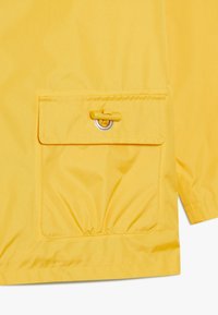 Sección de chaqueta impermeable amarilla con un bolsillo frontal con cierre de botón y dobladillo fruncido, fabricada con un material liso y texturizado.