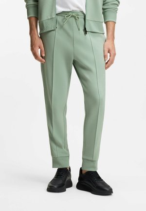 Uomo con pantaloni da jogging verde chiaro con cuciture frontali, polsini a coste, vita con coulisse e sneakers nere, in piedi davanti a uno sfondo bianco.