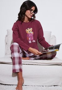 Bordó hosszú ujjú felső maci grafikával; bordó, fehér és szürke kockás pizsamanadrág; modell magazint olvas a kanapén.