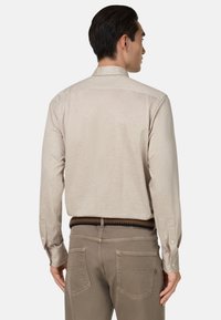 Beżowa koszula z długim rękawem i kołnierzykiem, gładka w dotyku, z zapięciem na guziki przy mankietach, noszona z ciemnymi spodniami i plecionym paskiem.