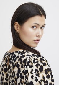 Leoparda raksta kleita bēšā un melnā krāsā ar teksturētu audumu, ar riņķveida atvērumu mugurā un pūkainām piedurknēm. Redzamas zelta auskari.