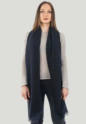 Vrouw met steil bruin haar draagt een lichtgrijze trui met lange mouwen, zwarte broek en een lange donkerblauwe sjaal die om haar nek is gedrapeerd.