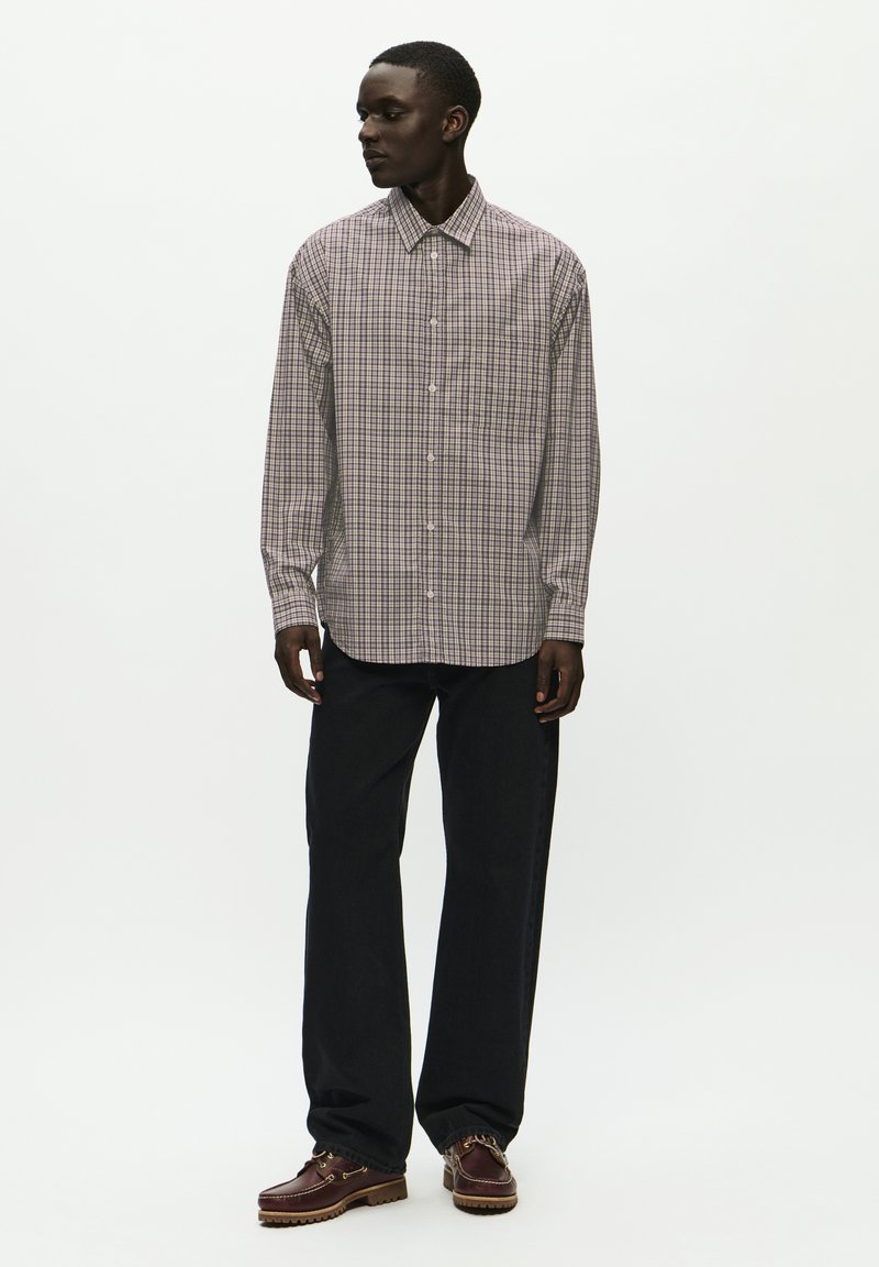 Camicia a maniche lunghe con bottoni, in un motivo a quadretti grigio e viola, indossata con jeans scuri e scarpe marroni. Sfondo neutro.
