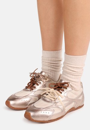 Μεταλλικά ασημί και μπεζ αθλητικά παπούτσια με ομαλή επιφάνεια και διάτρητο σχέδιο. Καφέ και μπεζ κορδόνια δεμένα μπροστά. Ριμπ λευκές κάλτσες.
