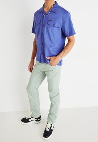 Kortärmad, knäppbar skjorta i livfull blå tyg; två bröstfickor. Kombinerad med ljusgröna byxor och svarta sneakers med vita sulor.