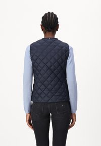 Marineblau gesteppte Weste mit Stehkragen, kombiniert mit einem hellblauen Langarmshirt. Schwarze Jeans runden das Outfit ab.