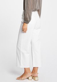 Jeans anchos y cortos de color blanco, fabricados en denim, con dos bolsillos traseros, combinados con una blusa texturizada de patrón marrón y sandalias de tacón bloque doradas con tiras.