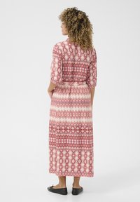 Abito maxi di colore rosa chiaro con motivi geometrici rossi, maniche a tre quarti, vita cintrata e tessuto fluente. Scarpe nere piatte indossate.