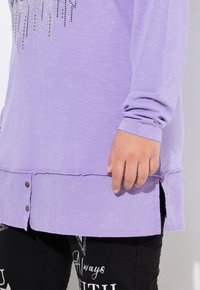Top lungo lavanda con orlo arrotondato, caratterizzato da un design di strass sul collo e due bottoni sulla cucitura laterale. Tessuto testurizzato.