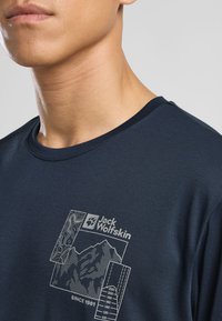 Marineblaue Kurzarm-T-Shirt aus glattem Material; mit einem weißen grafischen Logo und einem Bergdesign mit topografischen Details.