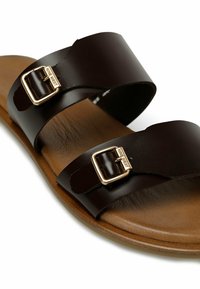 Brown leather sandal with two wide straps, each featuring a gold-tone buckle. Soft footbed, smooth texture, and a natural wood-tone sole.