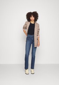 Blazer de cuadros en tonos marrón y negro sobre una camiseta negra, combinado con jeans azul de cintura alta y botas de tobillo color crema.