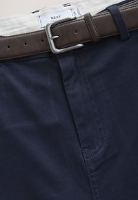 Navy blue trousers with a brown leather belt threaded through loops, showing the waist label inside the waistband.