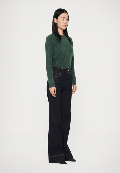 Woman with long black hair wearing a green sweater, high-waisted wide-leg dark jeans, and black pointed shoes, standing against white background.