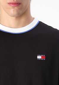 Läheltä kuva henkilöstä, joka käyttää mustaa Tommy Jeans -paitaa, jossa on valkoinen ja sininen ribbikaulus sekä logo-patch rinnassa.