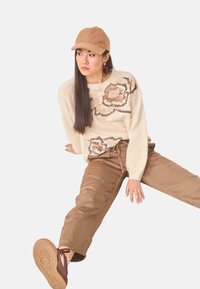 Joven mujer vestida con un suéter floral beige, pantalones marrones, gorra beige y zapatos marrones, sentada con una pierna extendida y mirando de lado.