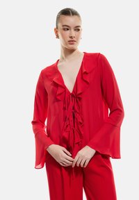 Blouse rouge transparente avec un col et des poignets froncés. Présente un décolleté en V plongeant et des détails de lien ajustables à l'avant. Texture lisse.