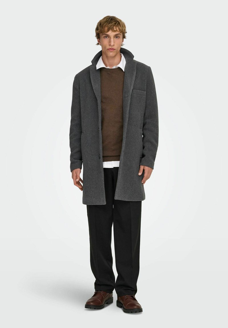 Manteau en mélange de laine gris avec col cranté, une poche poitrine simple, porté par-dessus un pull marron et une chemise blanche, associé à un pantalon noir et des chaussures marron.