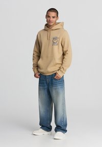 Felpa beige con cappuccio e design grafico sul petto, abbinata a jeans leggeri azzurri e sneakers bianche, in piedi davanti a uno sfondo neutro.