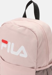 Mochila de tela rosa con una textura suave, que presenta el logo de Fila en blanco y rojo, un bolsillo de malla lateral y un asa de transporte negra.
