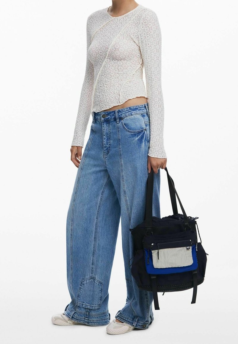 Femme portant un haut texturé blanc à manches longues et un jean bleu à jambes larges, portant un sac fourre-tout noir et bleu avec une poche avant.