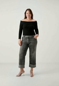 Γυναίκα που στέκεται, φορώντας μαύρο τοπ χωρίς ώμους, γκρι τζιν μαζεμένα και ασημένια ψηλοτάκουνα σανδάλια, μπροστά από απλό φωτεινό φόντο.