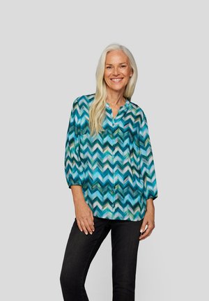 Bunt gemusterte Bluse mit Knopfleiste im blauen und grünen Zickzackdesign, mit 3/4-Ärmeln und lockerer Passform. Kombiniert mit dunklen Jeans.