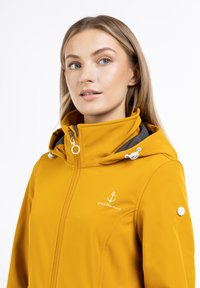 Gul vandtæt jakke med høj krave, grå indvendig foring, hvid ankerlogo og snøre hætte. Har lynlåslukning og detaljer i sidesømmen.