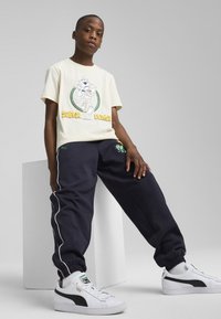 T-shirt crema con grafica cartone e testo "SUPER PUMA", abbinato a pantaloni della tuta blu navy con dettagli ricamati e piping bianco. Sneakers bianche.