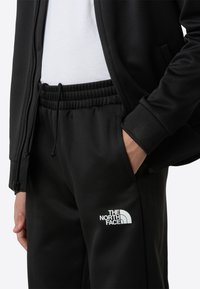 Čierny atletický tréningový oblek so zipom na bunde a elastickým pásom na nohaviciach. Na nohaviciach je logo a materiál má mäkkú textúru. Pod bundou je biele tričko.