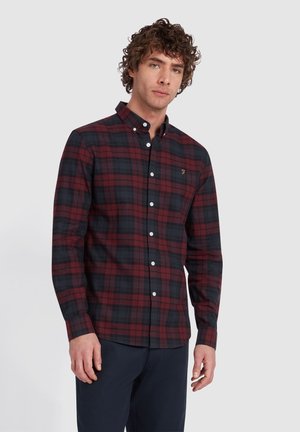 Hombre con pelo rizado que lleva una camisa de cuadros roja y negra abotonada y pantalones oscuros, de pie frente a un fondo claro y sencillo.