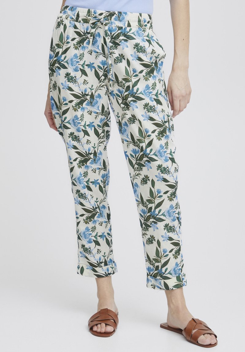 Blommiga byxor med krämfärgad botten och mönster av blå och gröna blommor. Elastiskt midjeband med dragsko. kanten är något uppvikt, matchat med bruna sandaler.