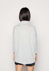 Donna con lunghi capelli scuri, che indossa un top a maniche lunghe grigio chiaro e pantaloni corti neri, vista da dietro su uno sfondo semplice.