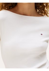 Weißes Baumwollhemd mit weitem Ausschnitt und kurzen Ärmeln, versehen mit einem kleinen gestickten Logo in Rot, Weiß und Blau auf der linken Seite.