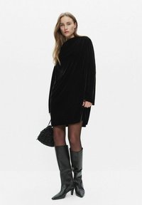 Schwarzes Samt-Minikleid mit langen Ärmeln, lockerer Passform und drapiertem Design. Kombiniert mit knielangen schwarzen Lederstiefeln und einer strukturierten schwarzen Tasche.