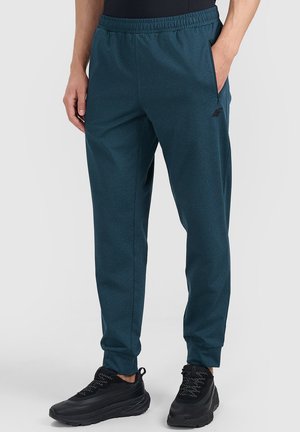Hombre con pantalones jogger de cintura elástica de color verde azulado oscuro y zapatos deportivos negros, con las manos descansando cerca de los bolsillos, de pie sobre un fondo liso.