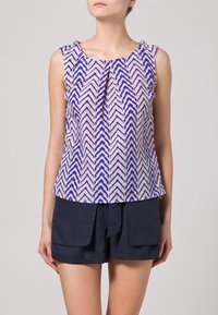 Bez rękawów bluzka w kremowym materiale z niebieskim wzorem w zygzak, z plisowanym dekoltem i luźnym krojem, noszona z ciemnymi szortami.